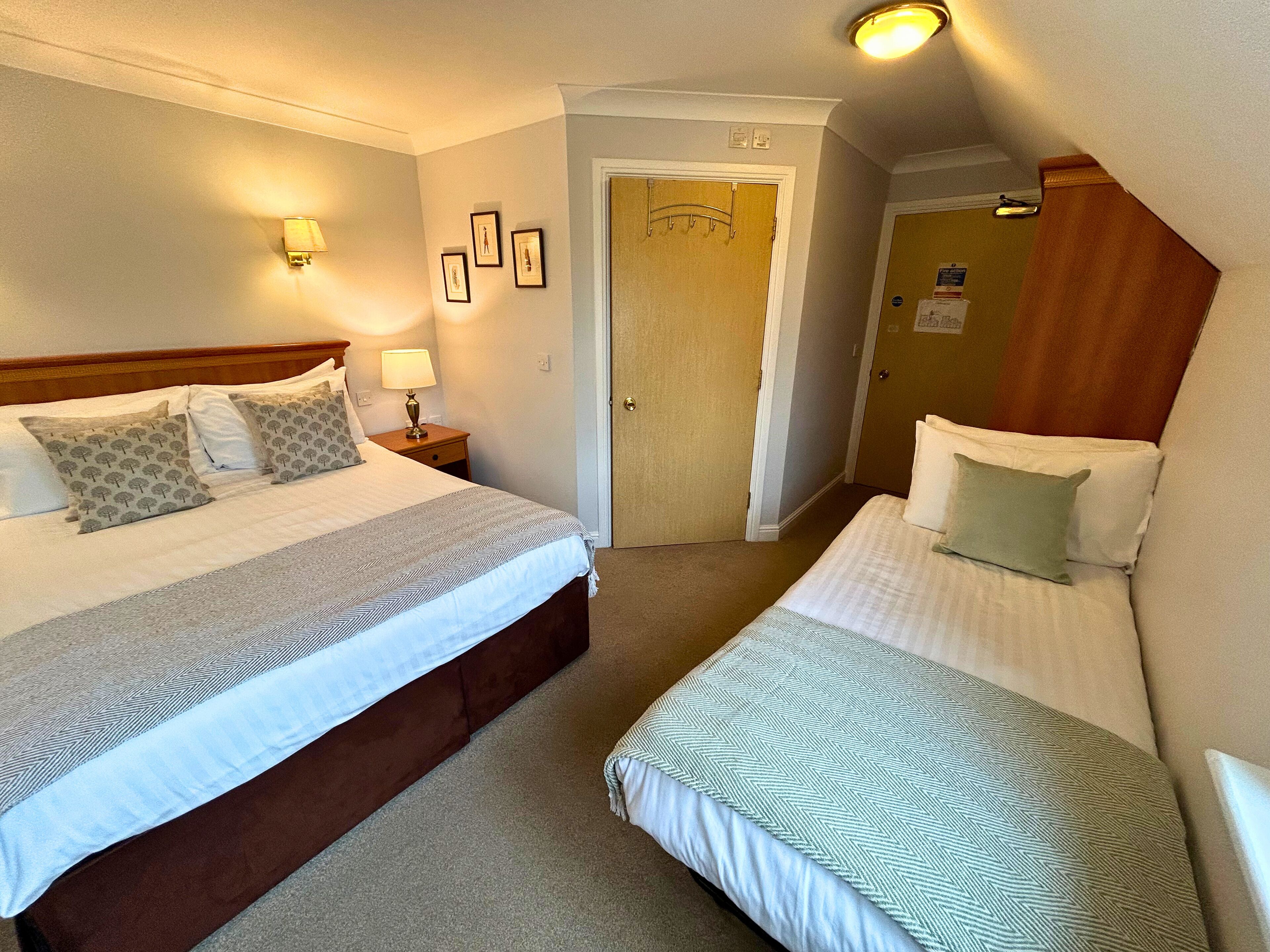 A hotel room with two beds, a nightstand with a lamp, a door with a coat rack, and a wall with framed pictures.
