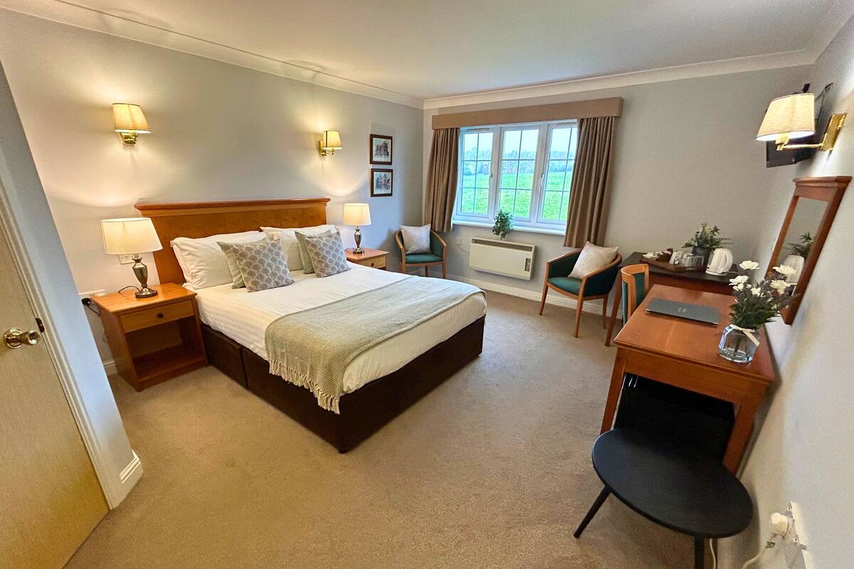 A hotel room with a bed, bedside tables, a desk, a chair, a window with curtains, and wall-mounted lamps.