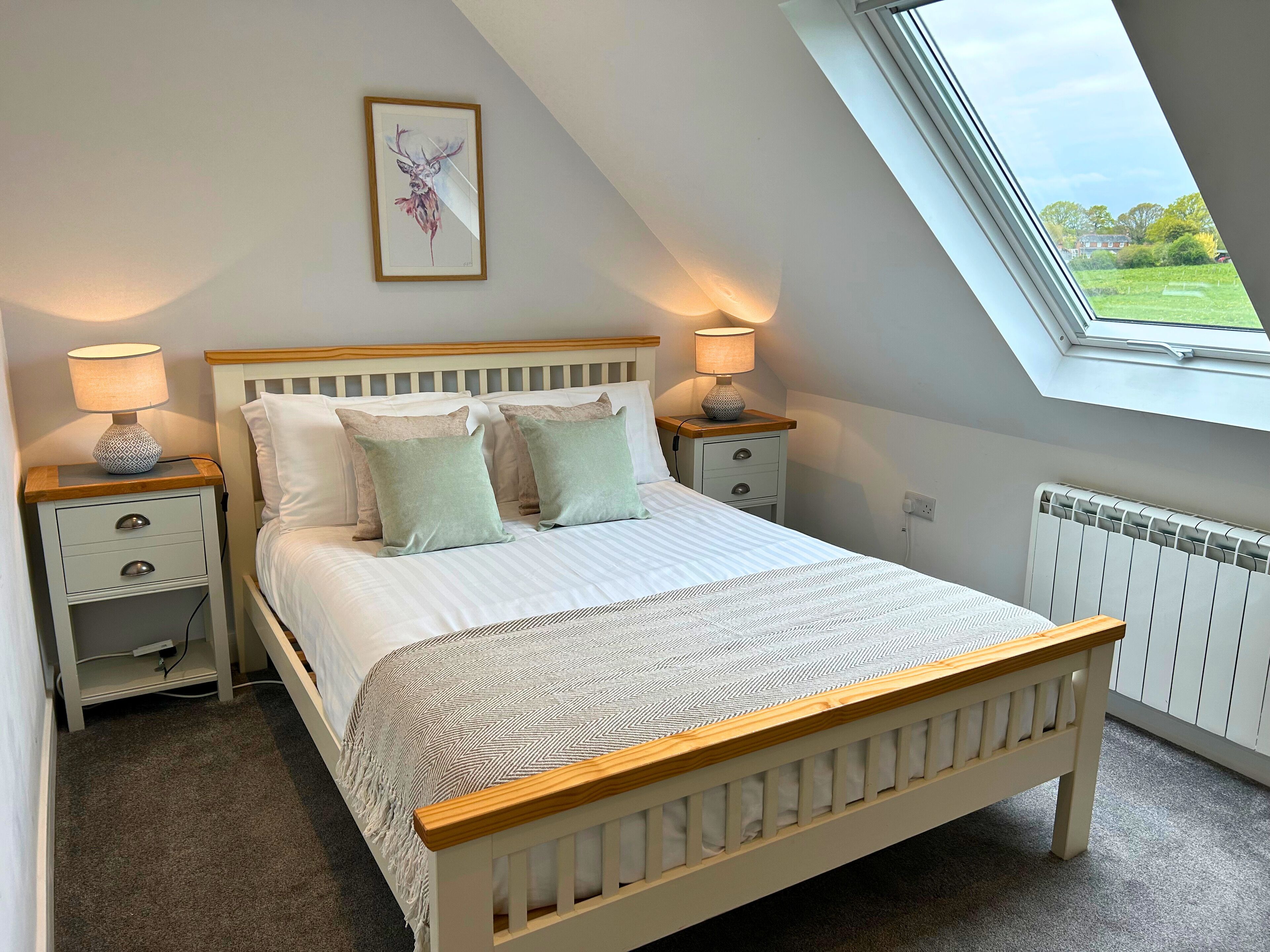Une chambre avec un plafond en pente, un lit doté d’une tête de lit en bois, deux tables de chevet avec des lampes, un tableau encadré au mur et une fenêtre donnant sur un paysage verdoyant.