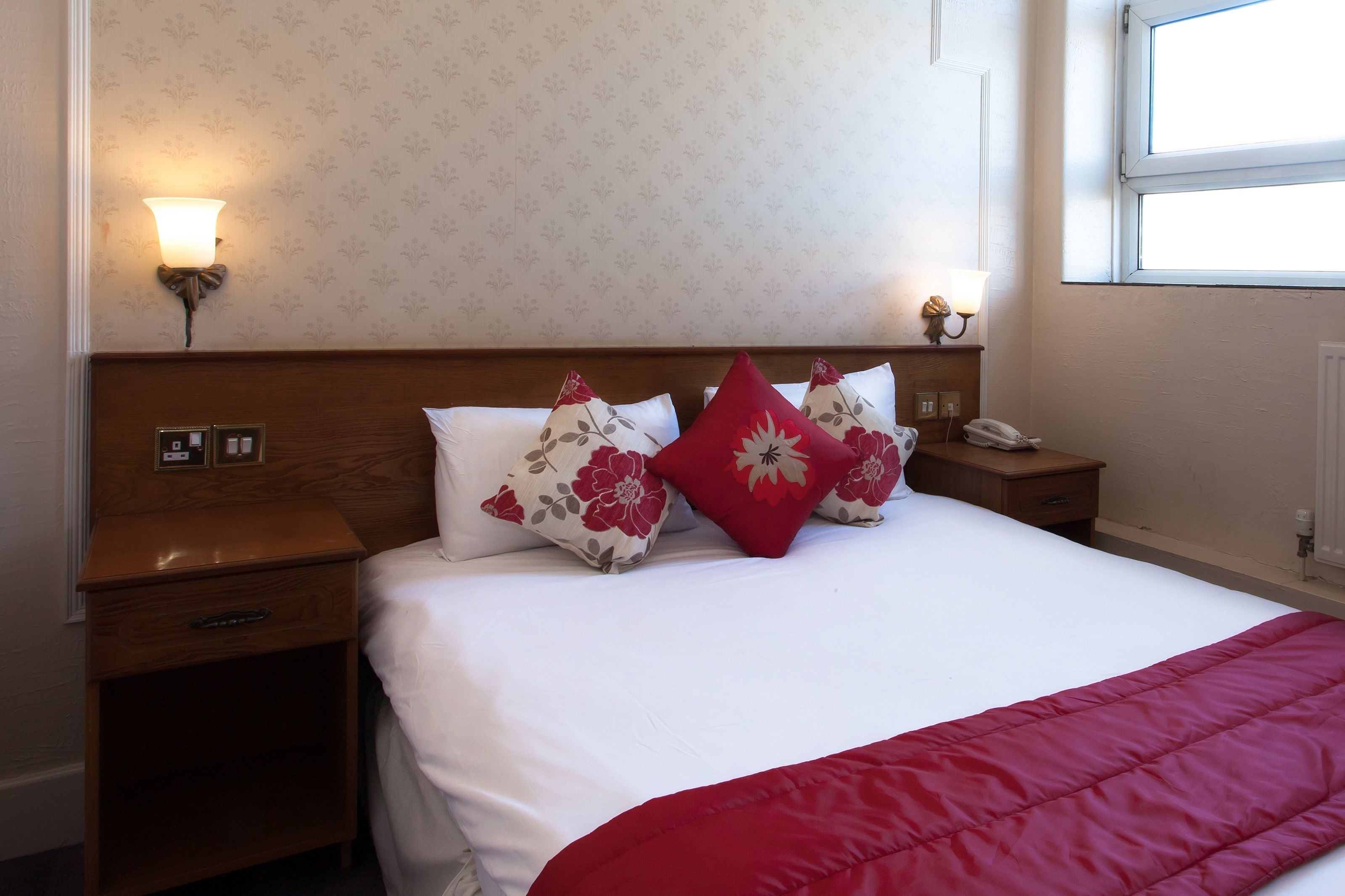 A neatly made bed with white bedding and red floral pillows, a wooden nightstand with a lamp, and a window with a view of the sky.