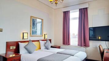 Habitación de hotel con una cama doble, mesitas de noche, televisión, ventana con cortinas, candelabro y un cuadro enmarcado en la pared.