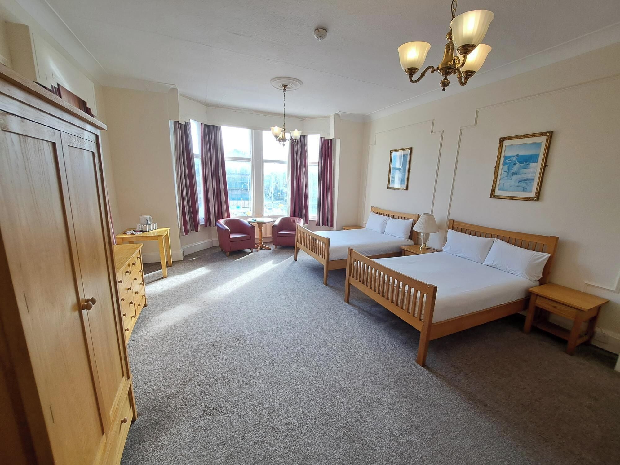 A hotel room with two single beds, a wooden wardrobe, a desk with a chair, and a window with curtains.