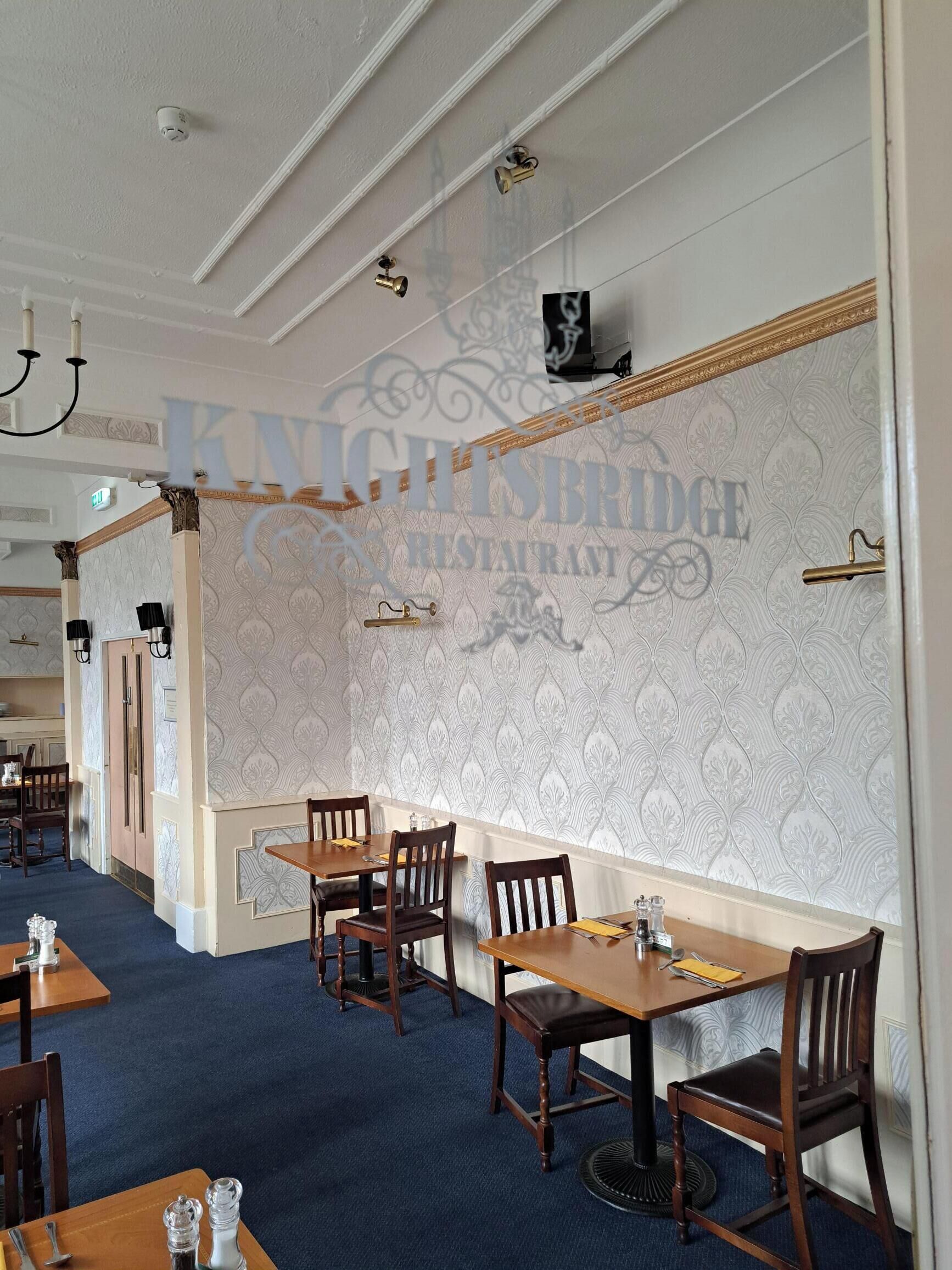 A restaurant interior with patterned wallpaper, wooden tables and chairs, and a chandelier.