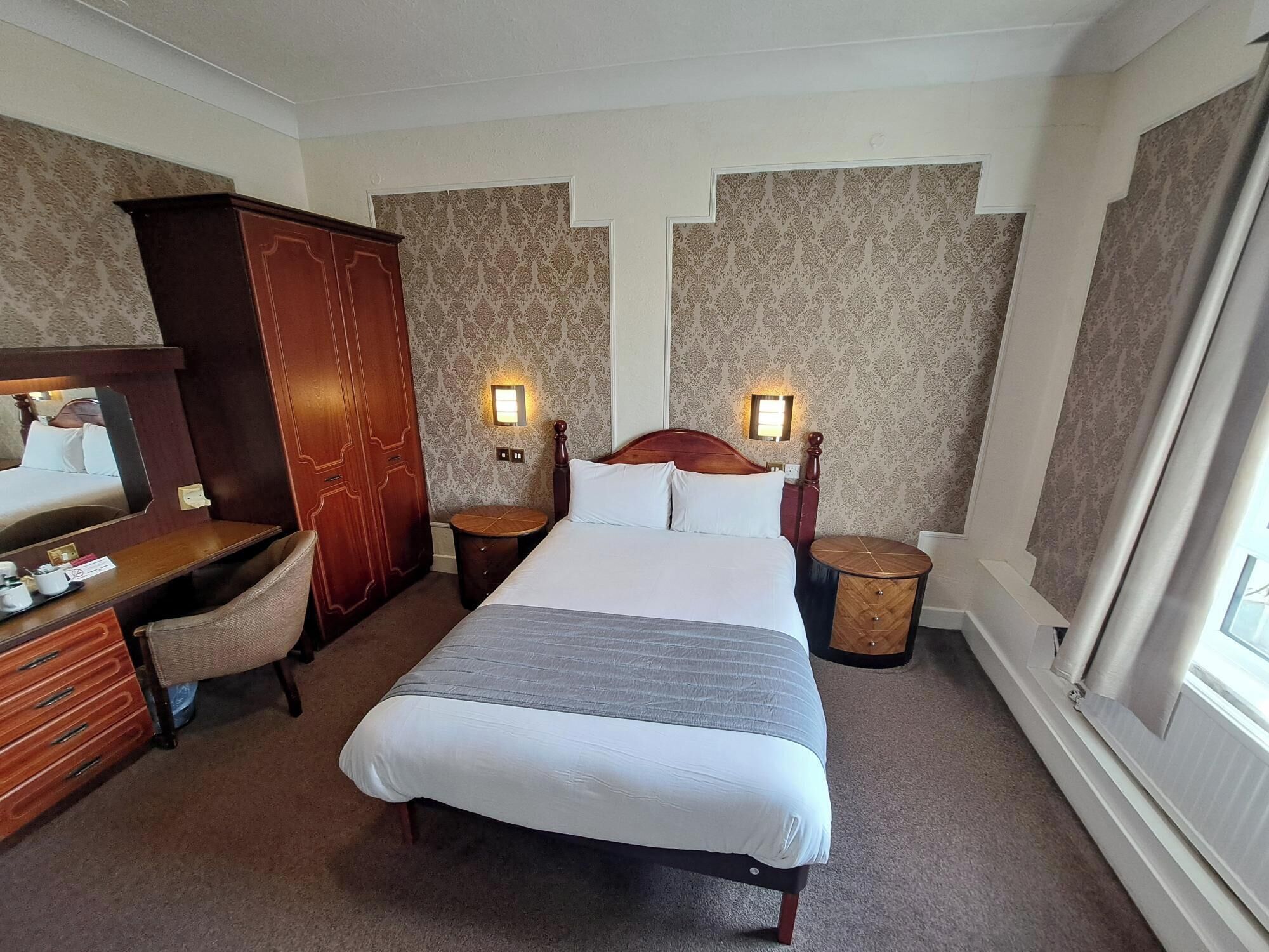 A hotel room with a large bed, a wooden wardrobe, a desk with a chair, and two bedside tables with lamps.