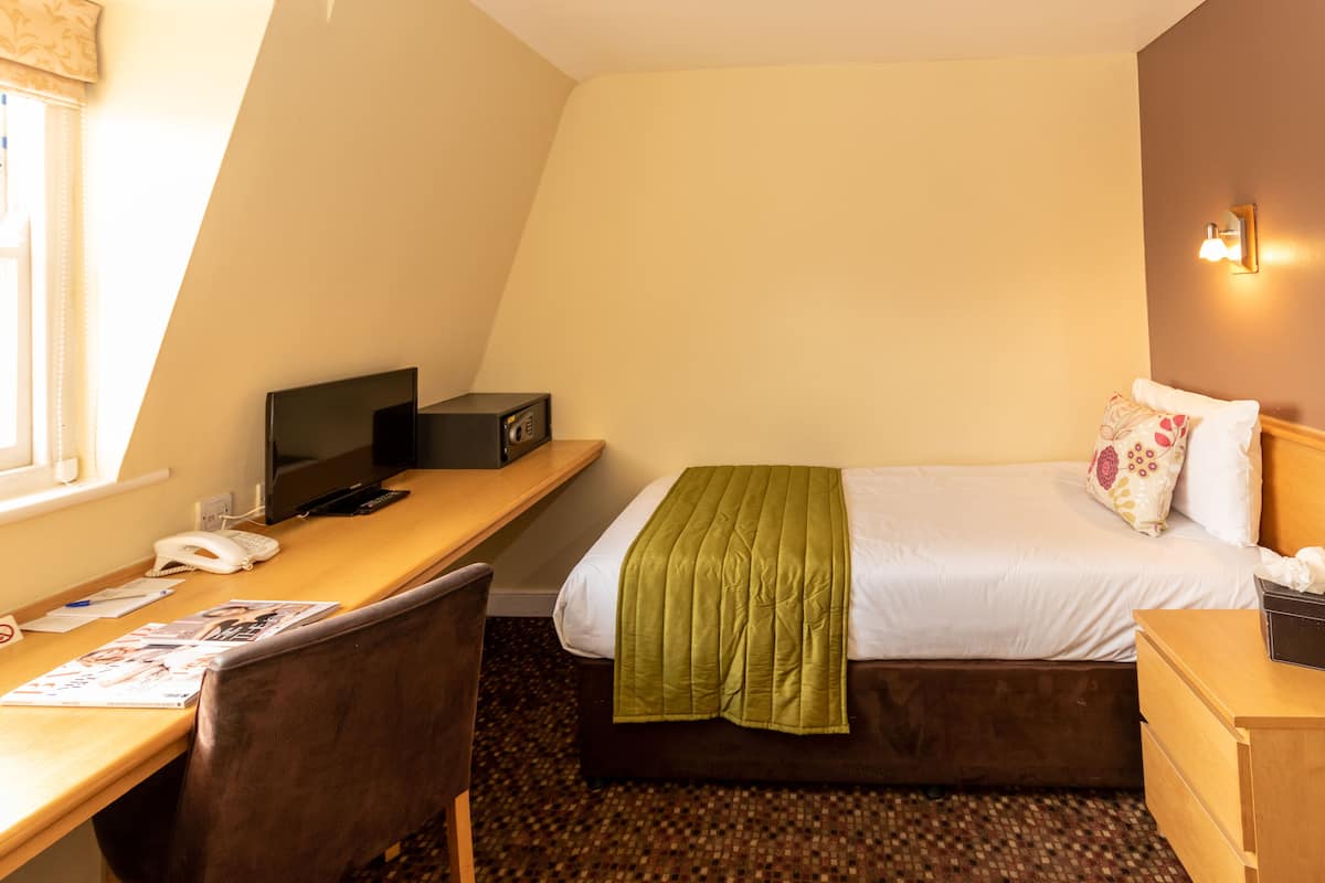 A hotel room with a bed, a desk with a computer, a chair, a telephone, and a bedside table.