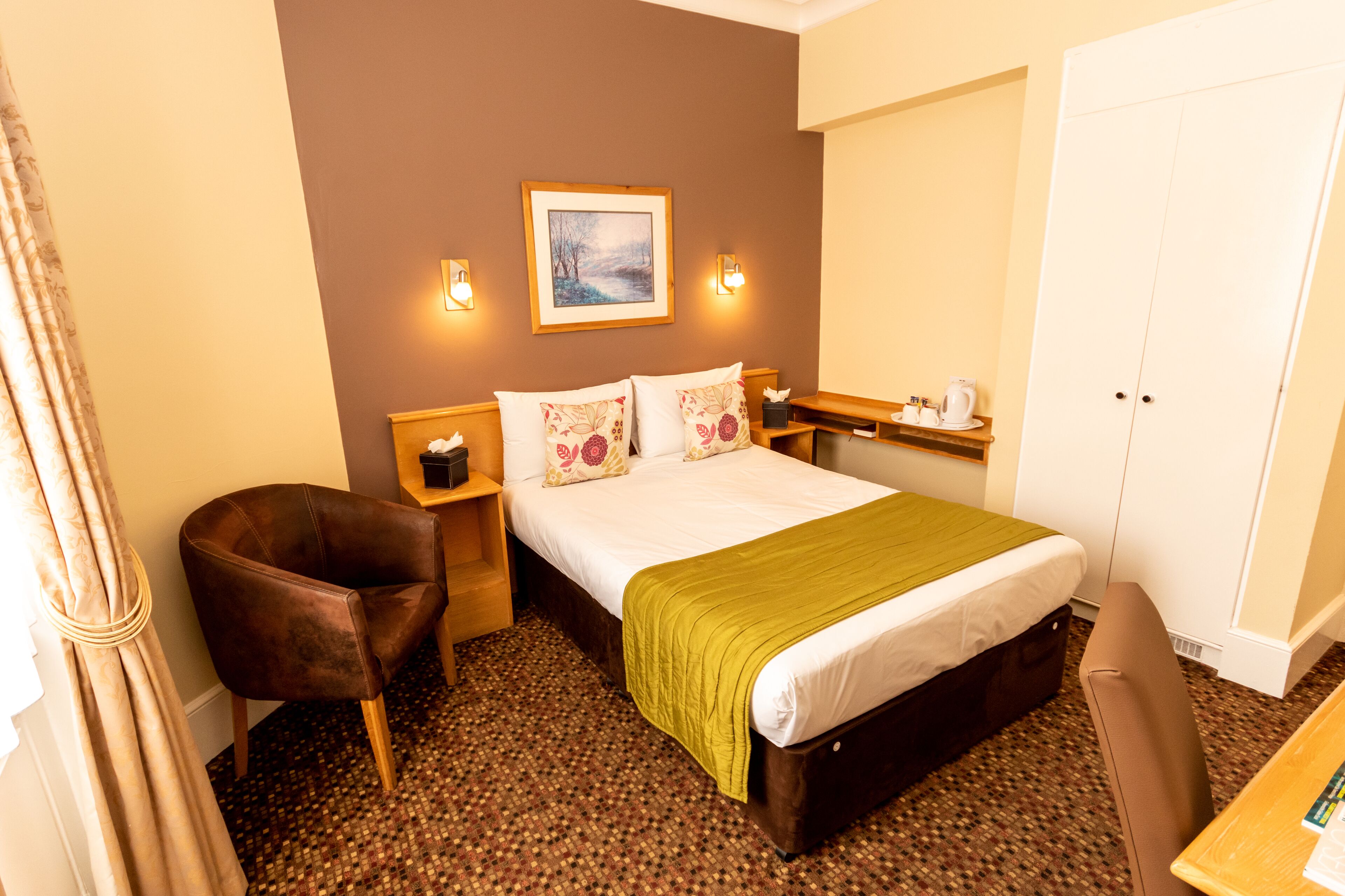 A hotel room with a bed, a chair, a desk, a wardrobe, and a painting on the wall.