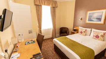 A hotel room with a bed, a desk with a computer, a chair, a TV, and a window with curtains.