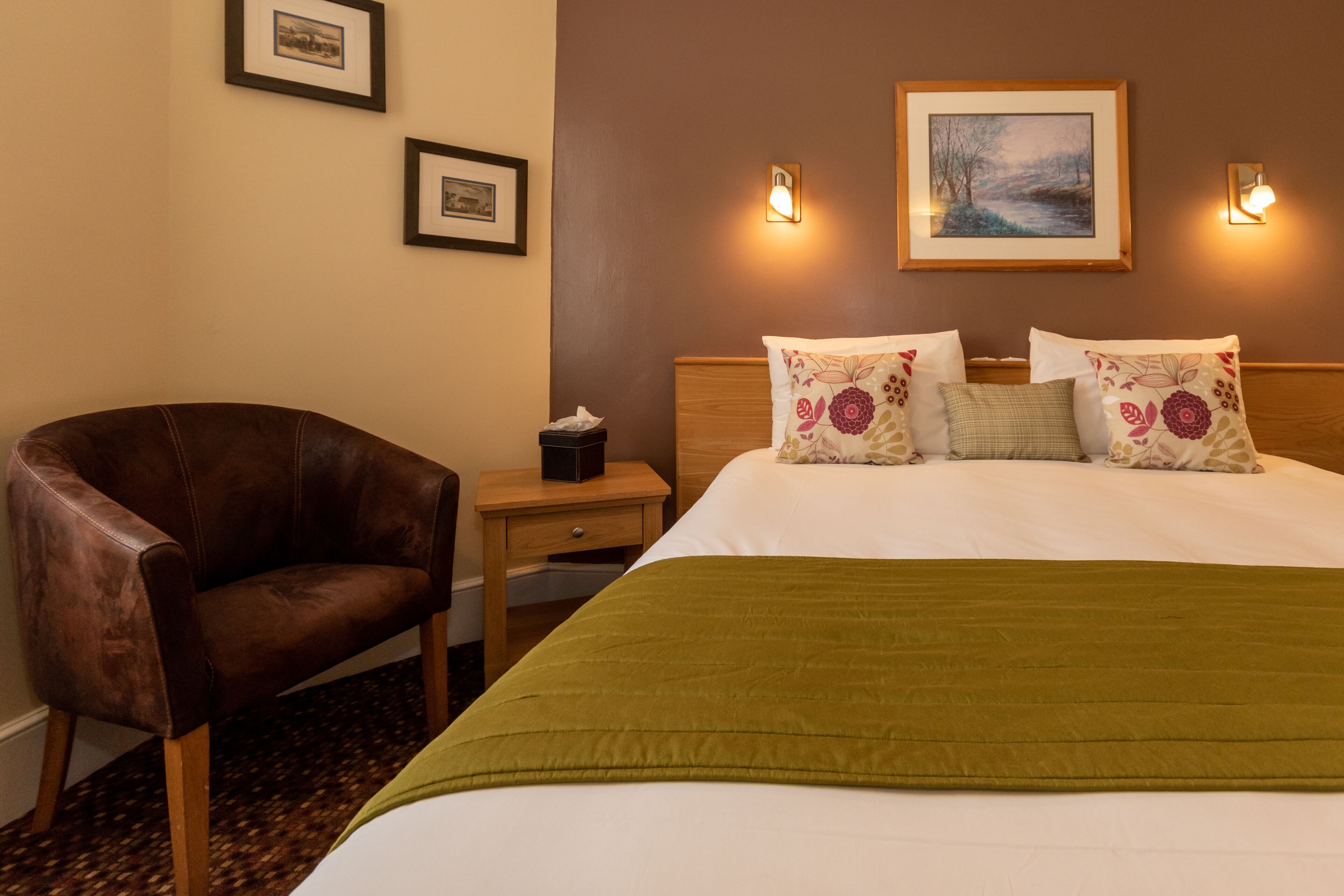A hotel room with a bed, a chair, a nightstand, and framed pictures on the wall.