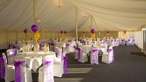 Un espacio para eventos con carpas, mesas y sillas, decorado con tela blanca y púrpura.