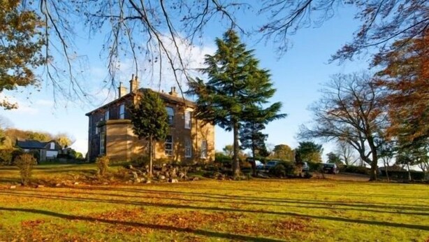 Una casa de dos pisos con fachada de piedra, rodeada por un césped cuidado y árboles.