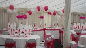 Un espacio para eventos con carpas, mesas redondas, sillas blancas con cintas rojas y globos rosa y blancos.