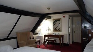A room with a sloped ceiling, a bed, a chair, a small table with a lamp, and a mirror on the wall. - The Falcon Hotel (Bromyard)