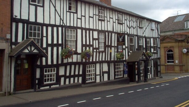 A traditional half-timbered building with a prominent chimney, a small porch with hanging flower baskets, and a signboard. - The Falcon Hotel (Bromyard)