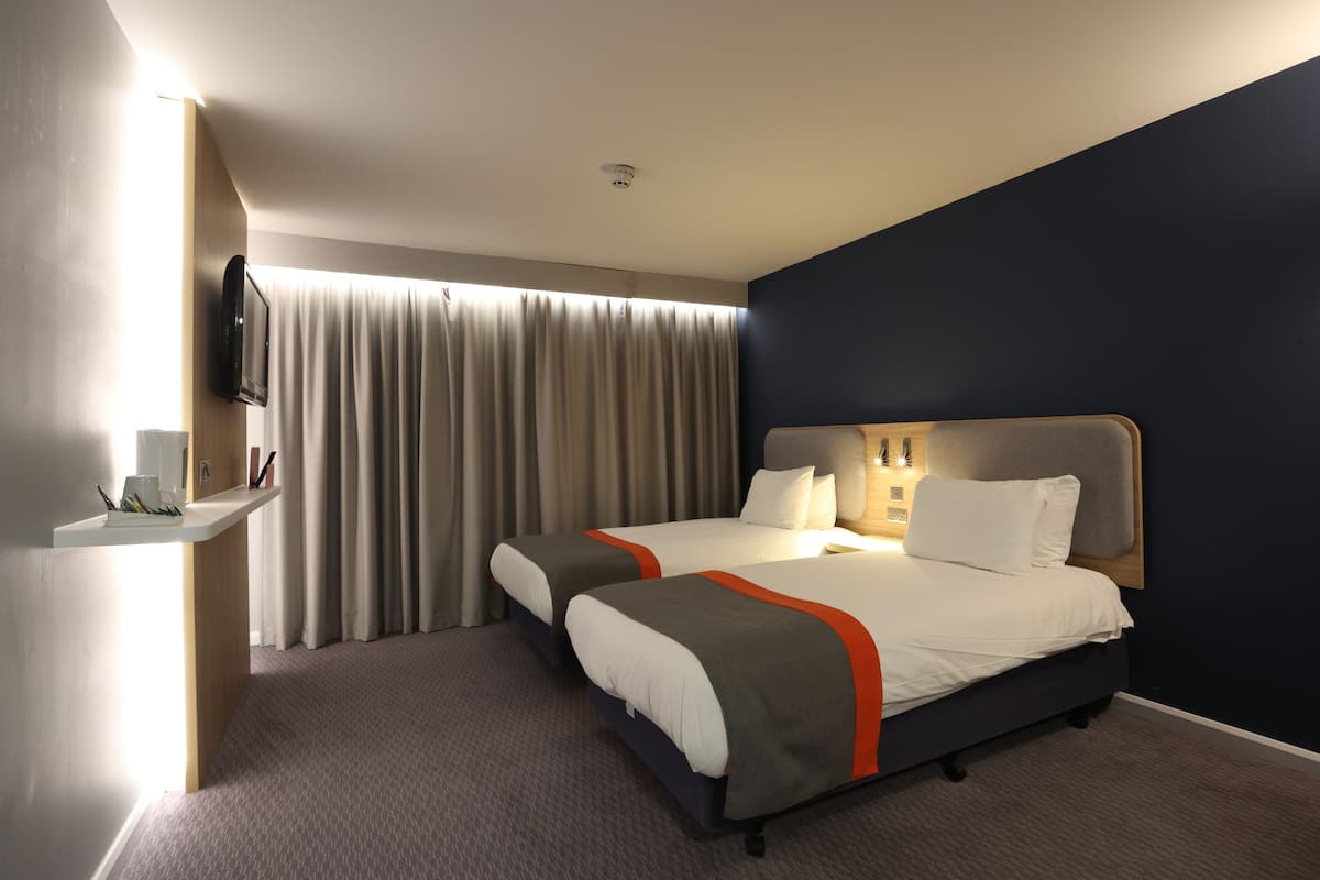 A hotel room with two beds, a desk, a TV, and a large window with curtains.