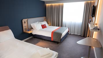 Habitación de hotel con una cama grande, una cama más pequeña, una mesita de noche con lámpara, un televisor montado en la pared y una ventana con cortinas.