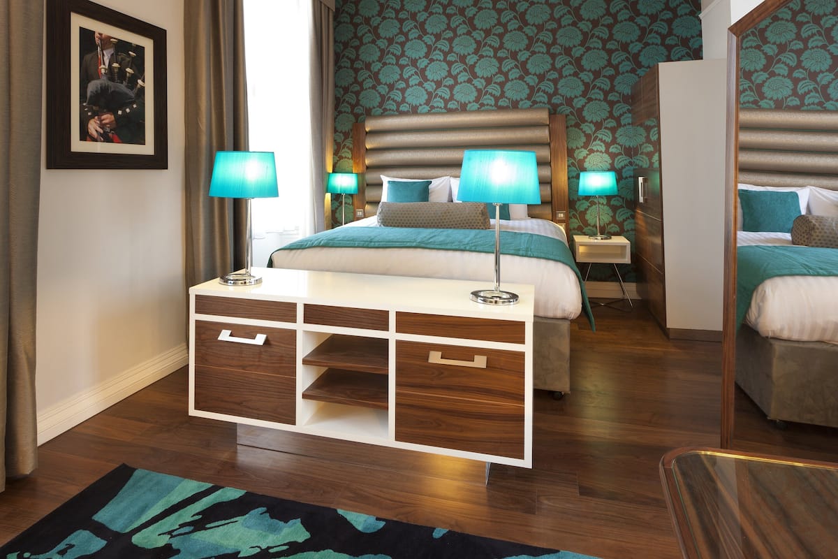 A hotel room with a large bed, two bedside tables with lamps, a wooden nightstand, a dresser with drawers, and a patterned wallpaper.