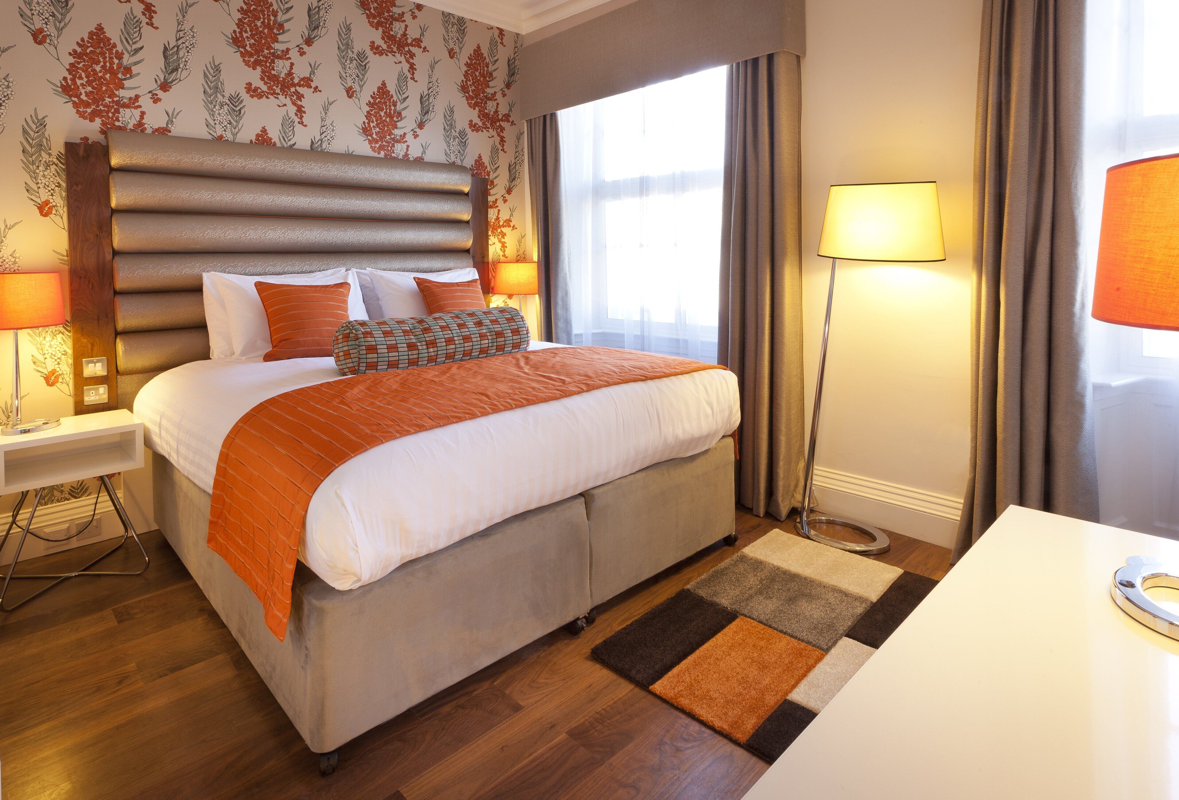 A hotel room with a large bed, a nightstand with a lamp, a desk with a lamp, and a window with curtains.