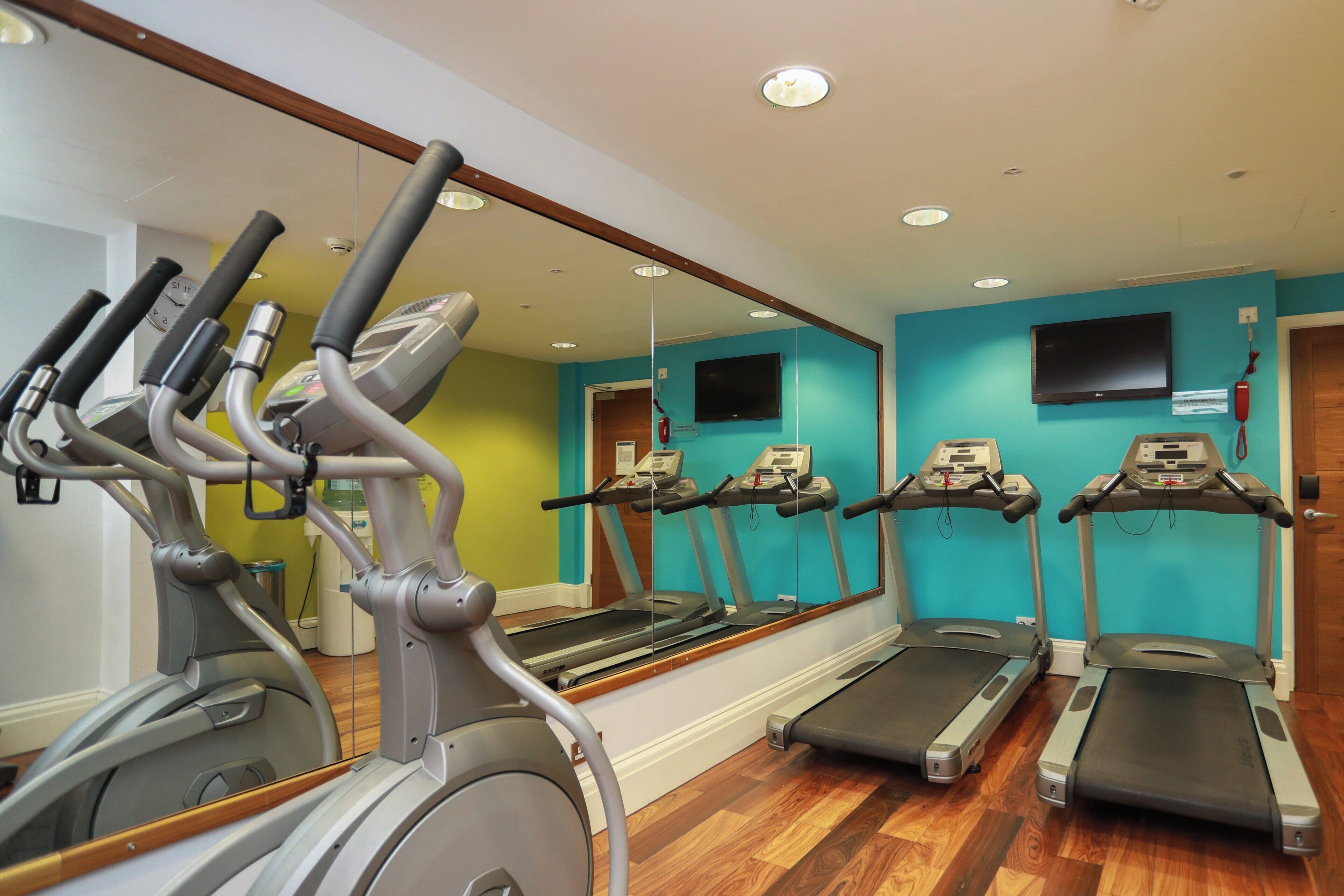 A gym with multiple treadmills and stationary bikes, wooden flooring, and a large mirror reflecting the equipment.