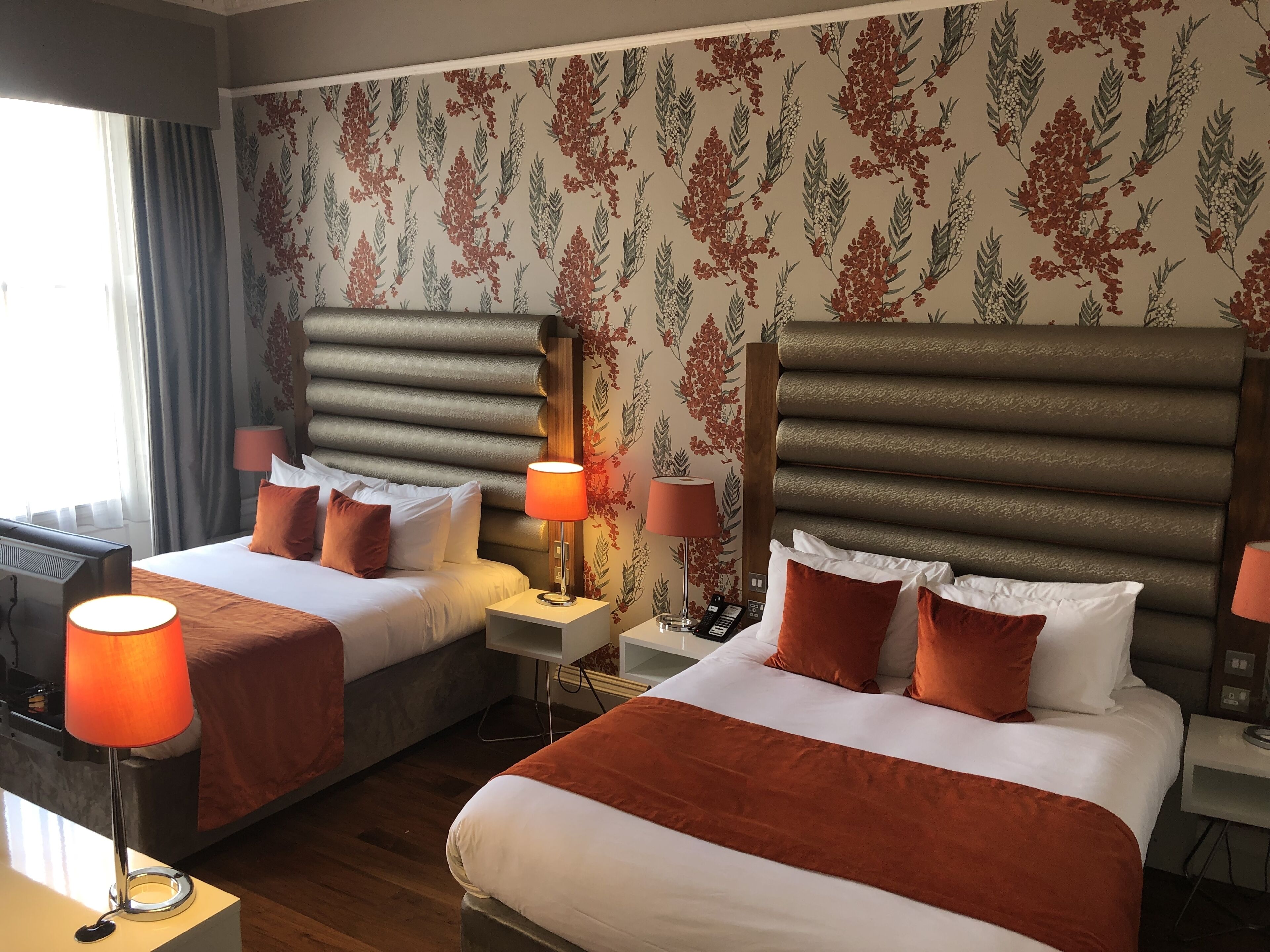 A hotel room with two beds, each with a red and white bedspread, and matching red lamps. The room has a wooden floor and a floral wallpaper design.