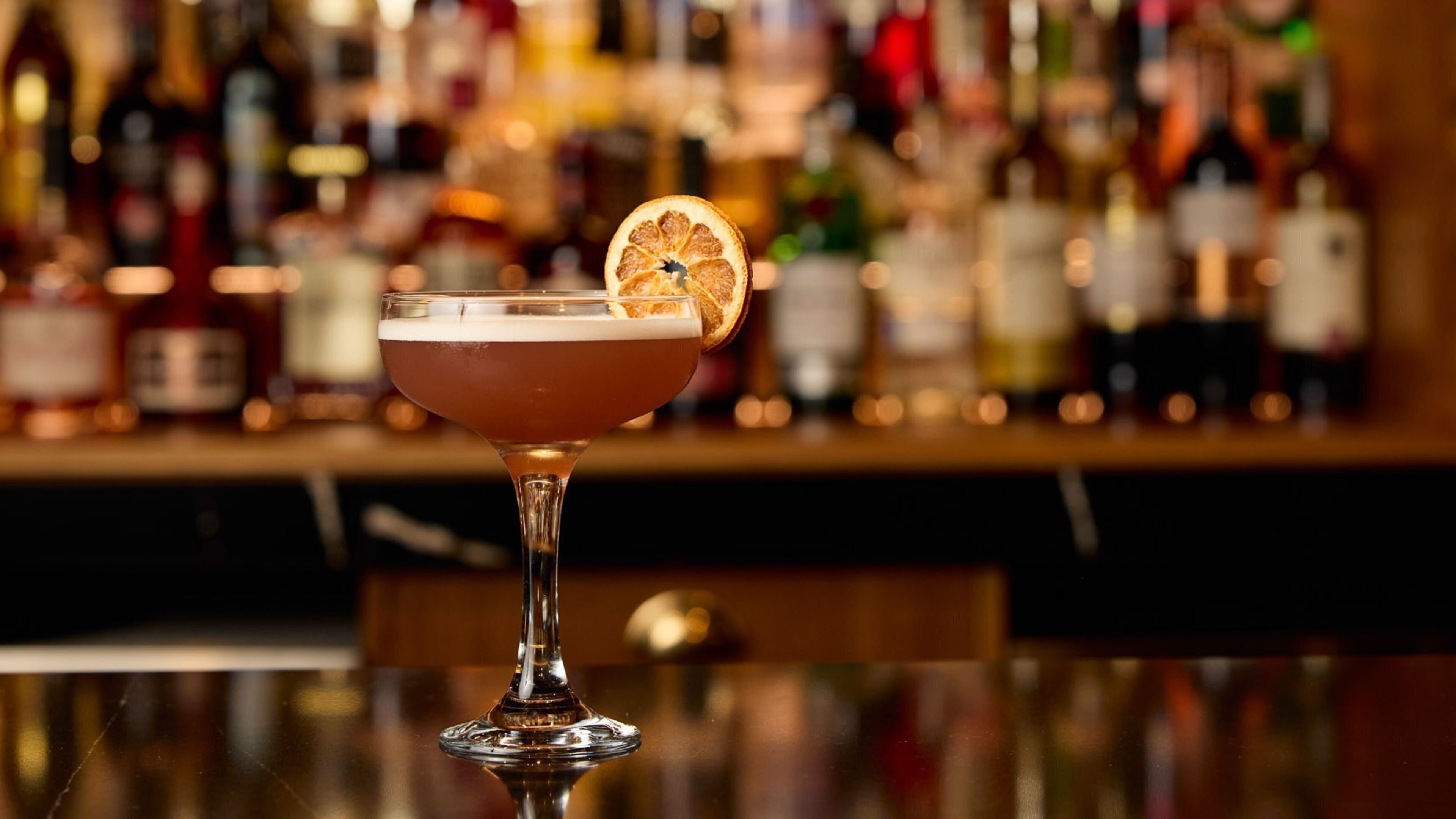 A cocktail in a stemmed glass with a slice of orange garnish on a bar counter.
