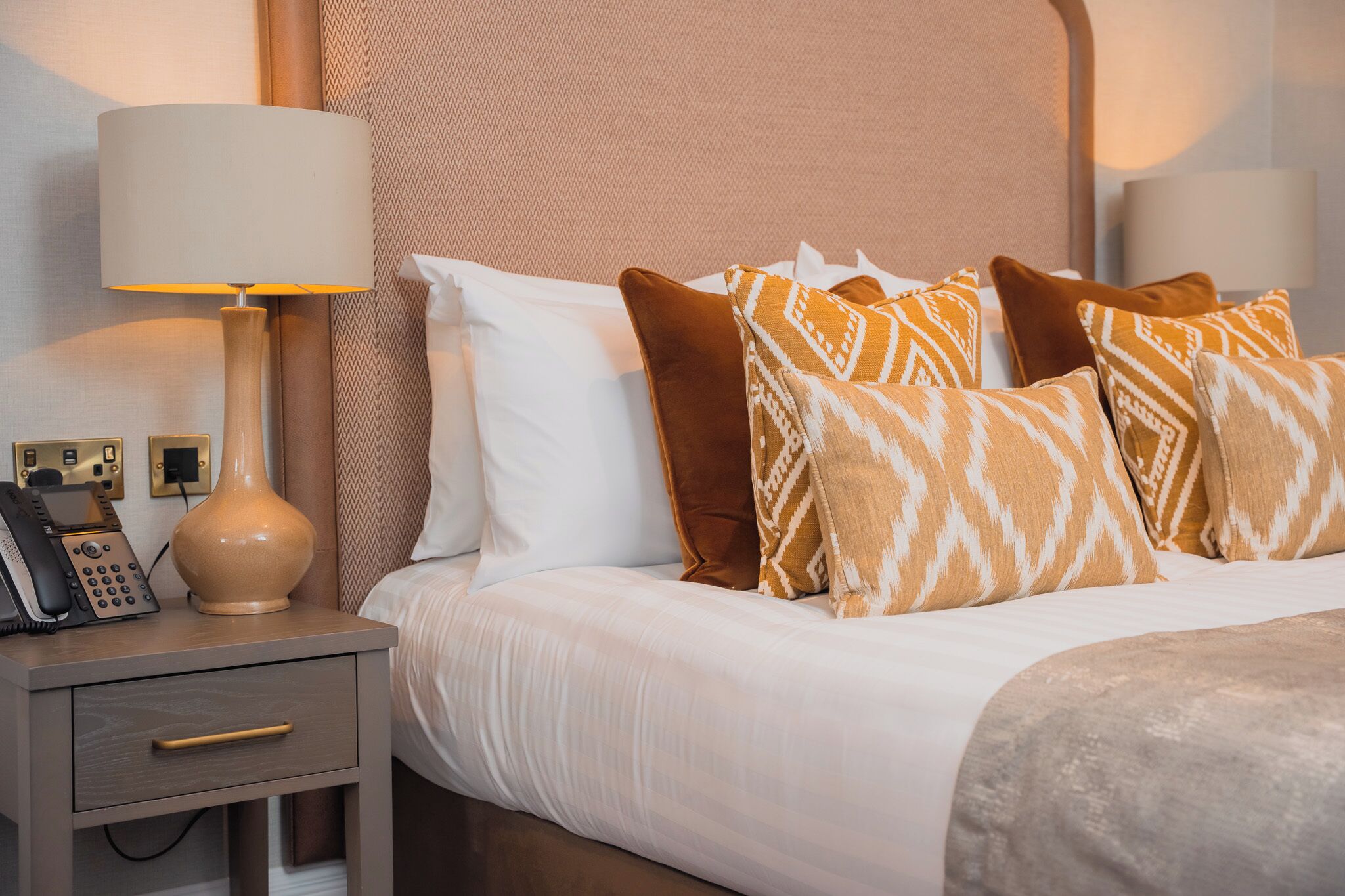 Een net opgemaakt bed met gekleurde kussens, een bedlampje en een nachtkastje met een telefoon en een lade.