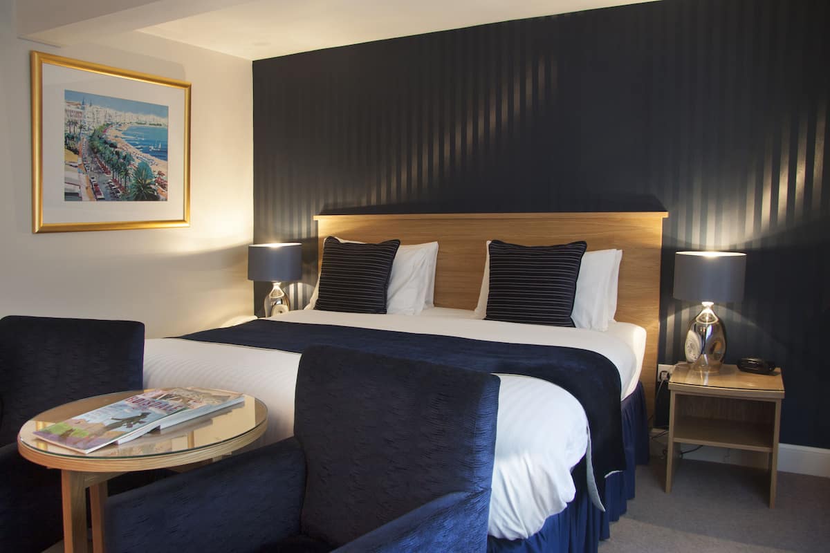 A hotel room with a large bed, two bedside lamps, a round table with a magazine, and a framed picture on the wall.