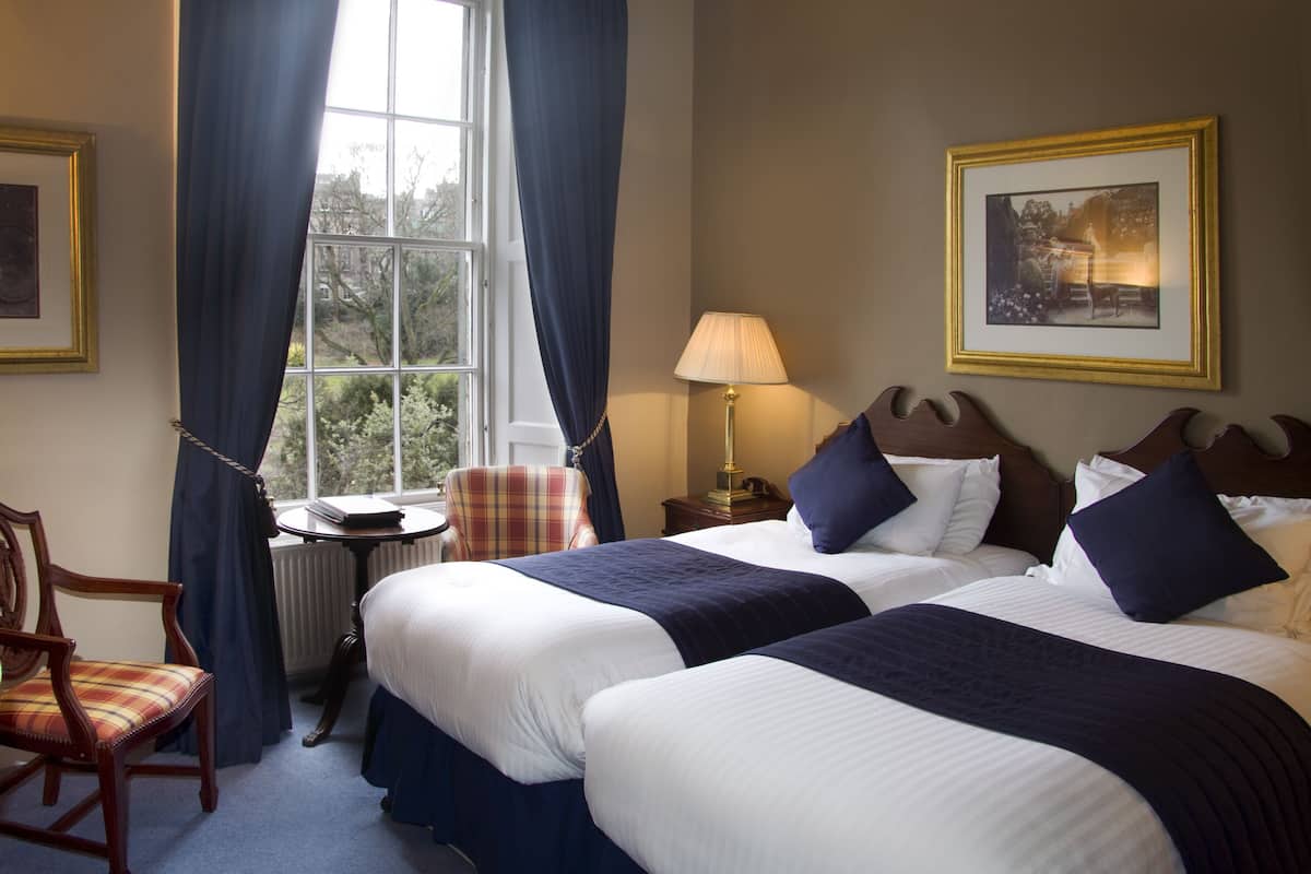 A hotel room with two beds, a chair, a table, a lamp, and a window with curtains.