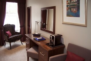 A hotel room with a desk, a chair, a coffee machine, a mirror, and a painting on the wall.