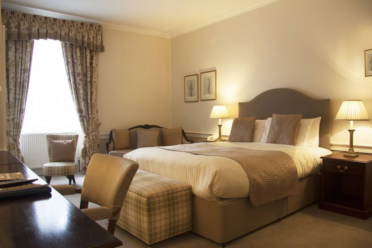 A hotel room with a large bed, two bedside lamps, a chair, a small table, and a window with curtains.
