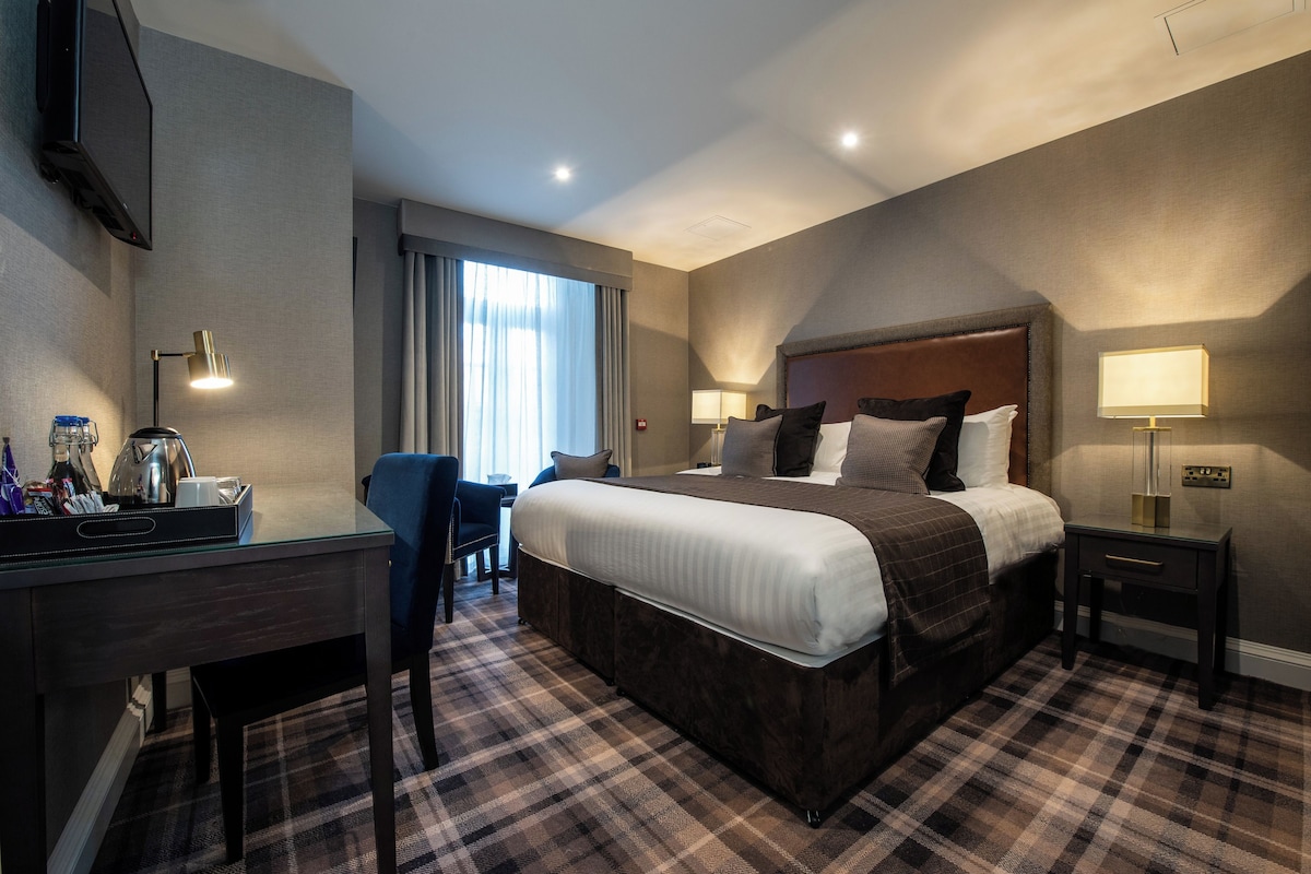Une chambre d’hôtel avec un grand lit, un bureau avec une chaise, un téléviseur fixé au mur et une fenêtre avec des rideaux.