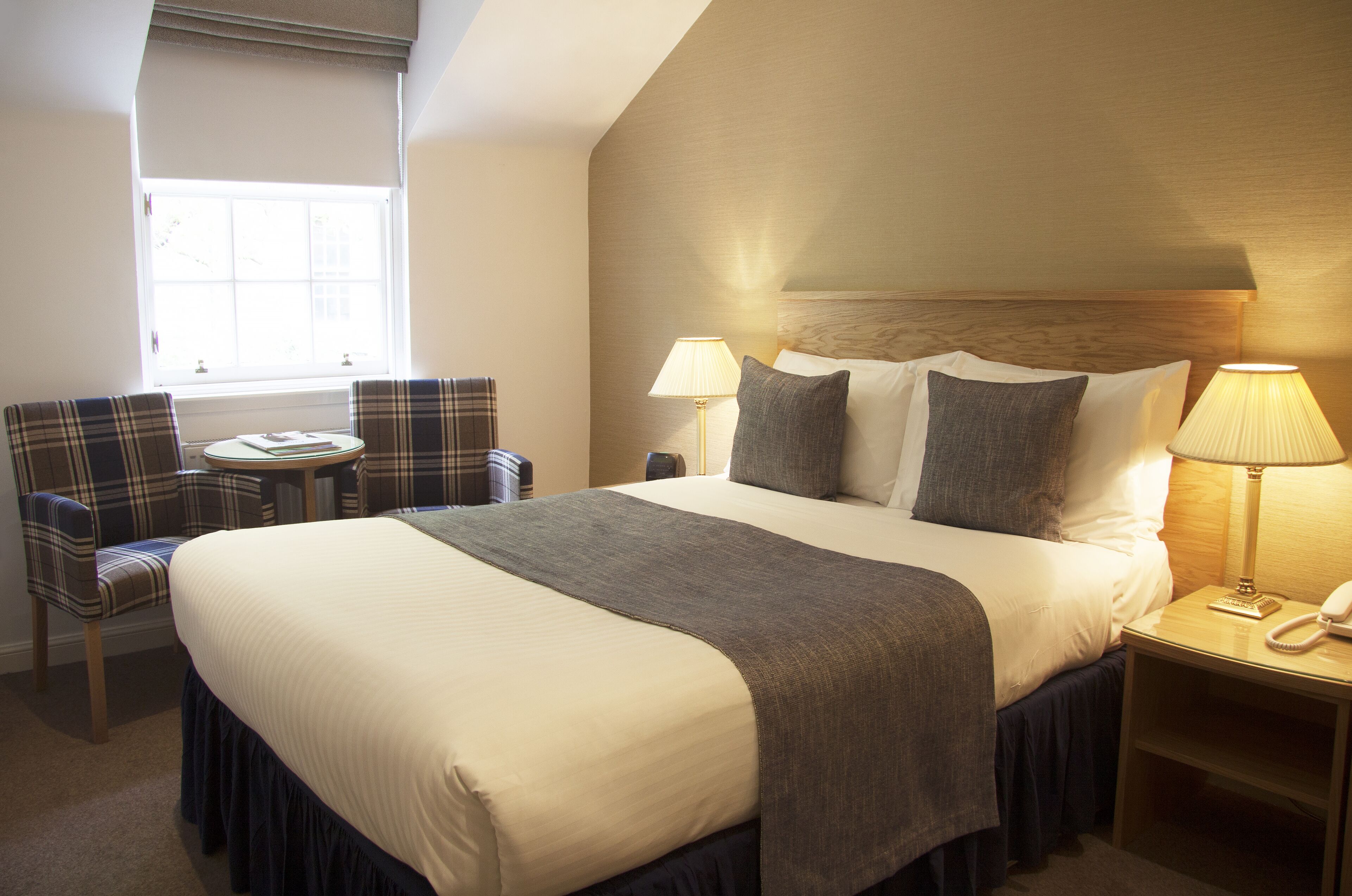 A hotel room with a large bed, two bedside lamps, a small table with a chair, and a window.