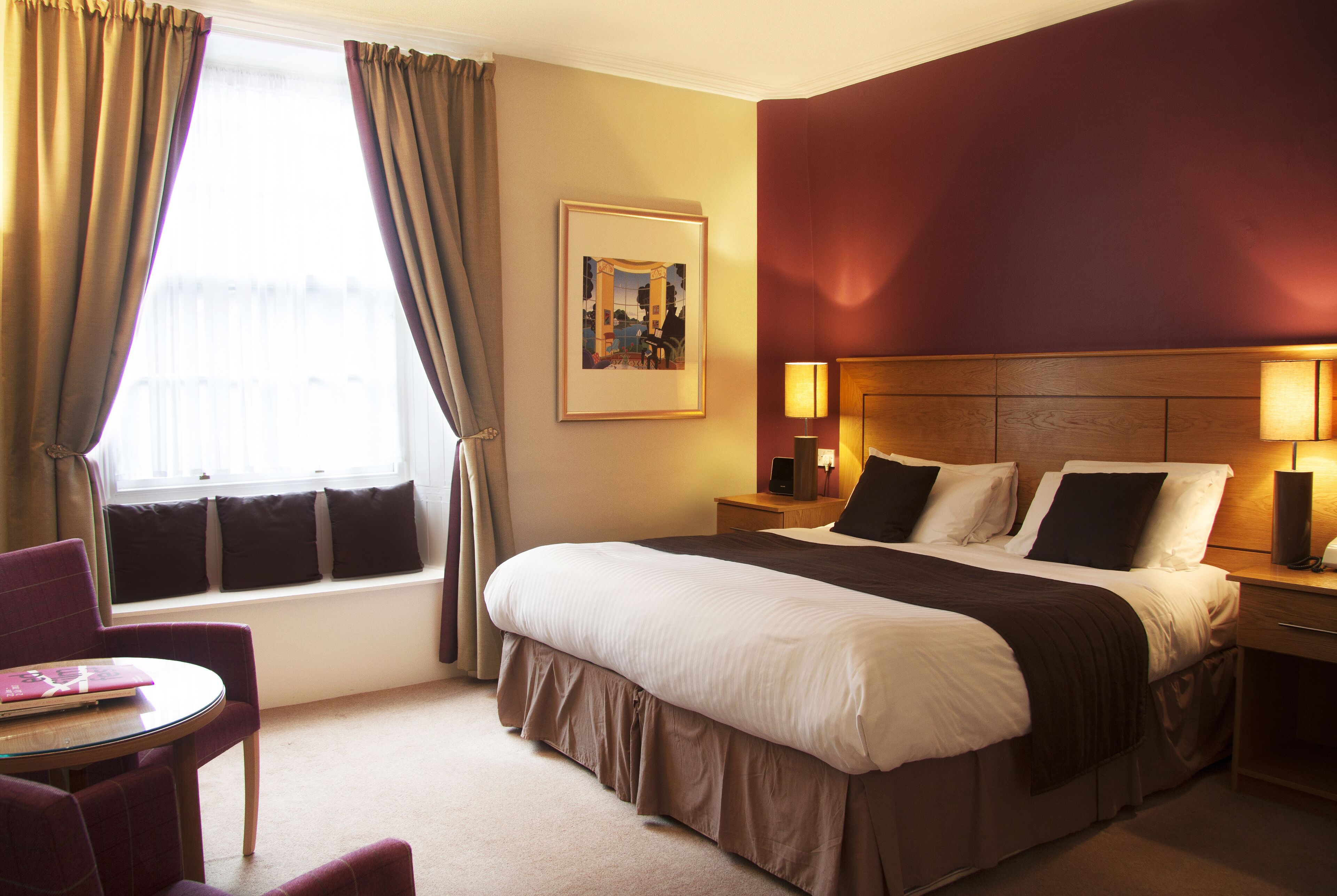 A hotel room with a large bed, a bedside table with lamps, a window with curtains, a small table with a chair, and a framed picture on the wall.