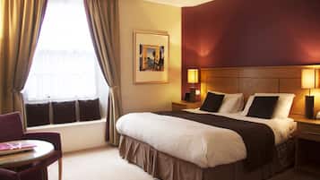 A hotel room with a large bed, a bedside table with lamps, a window with curtains, a small table with a chair, and a framed picture on the wall.