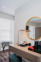 A hotel room with a desk, a round mirror, a lamp, a coffee maker, and a chair.