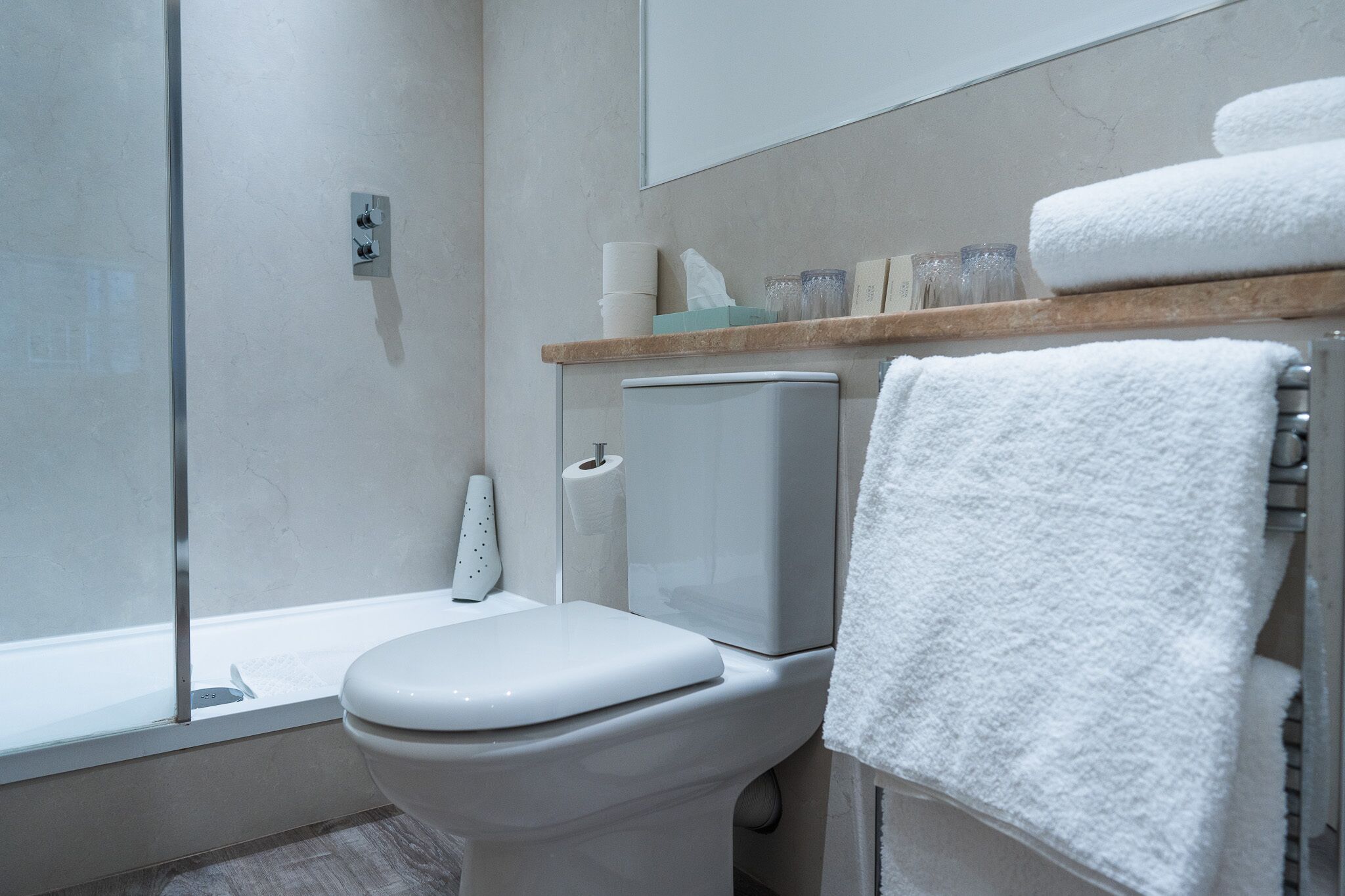 Un baño moderno con inodoro, cabina de ducha de vidrio, toallero con una toalla blanca y repisa de madera con artículos de aseo.