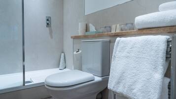 Un baño moderno con inodoro, cabina de ducha de vidrio, toallero con una toalla blanca y repisa de madera con artículos de aseo.