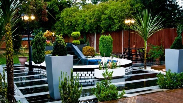 A modern outdoor patio with a circular water feature, wooden deck, and various plants. - Skylark B&B (Hounslow)
