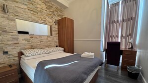 A modern bedroom with a stone wall, a wooden wardrobe, a desk with a lamp, and a bed with a grey blanket. - Skylark B&B (Hounslow)