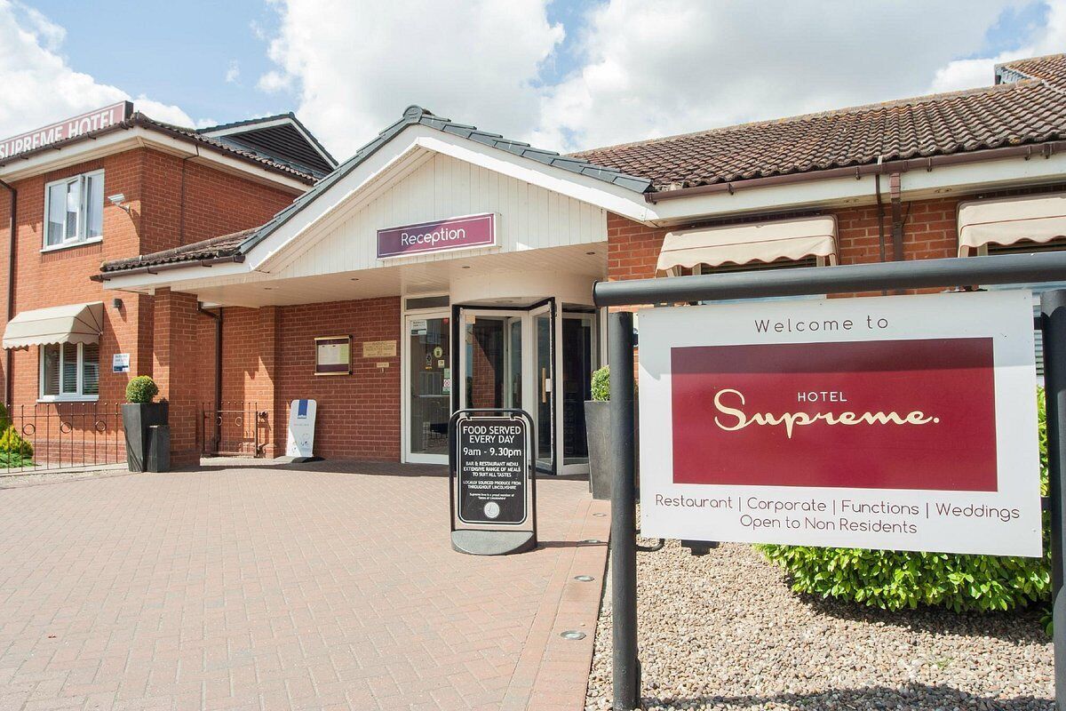 Een hotelingang met een receptiebalie, een bord waarop de openingstijden van de horeca staan vermeld, en een welkomstbord voor HOTEL Supreme.