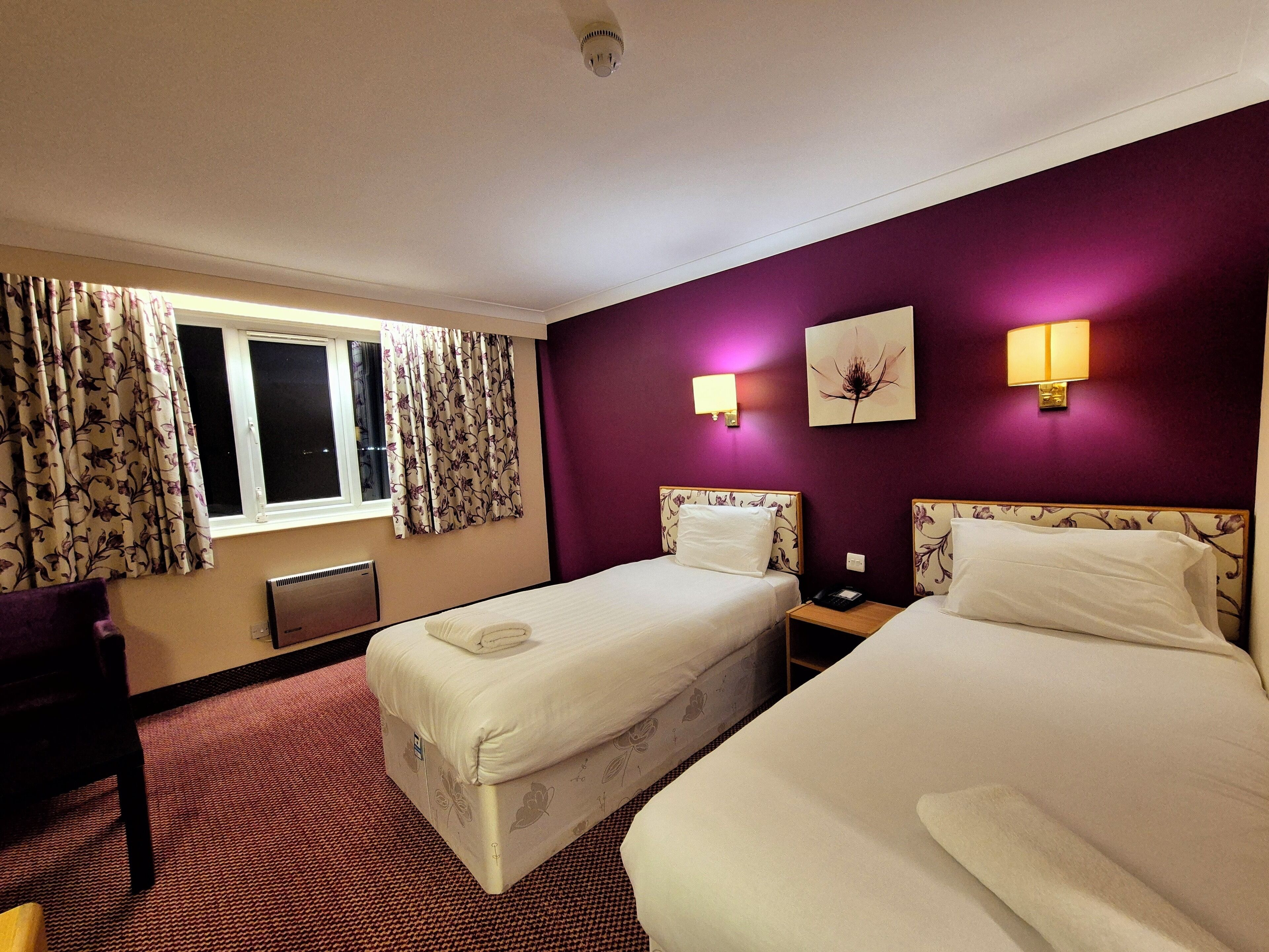 Une chambre d’hôtel avec deux lits, une fenêtre avec des rideaux, une lampe fixée au mur et un radiateur.