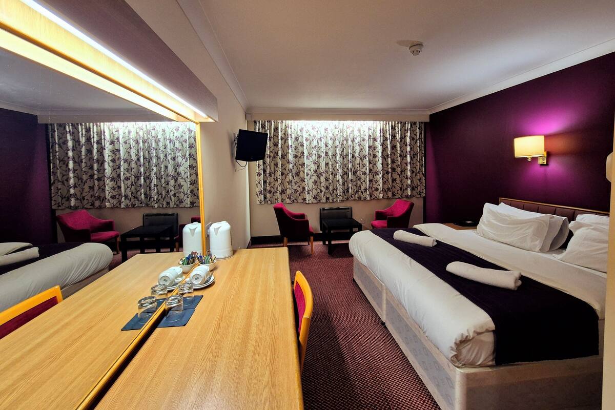 A hotel room with a bed, a desk with a chair, a table with a chair, a TV, and a window with curtains.