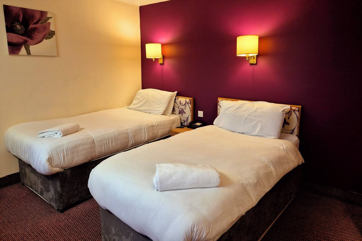 Two single beds with white linens and towels, a purple wall, and two wall-mounted lamps.