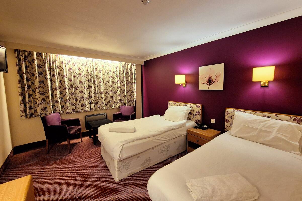 A hotel room with two beds, a desk, a chair, and a window with floral curtains.