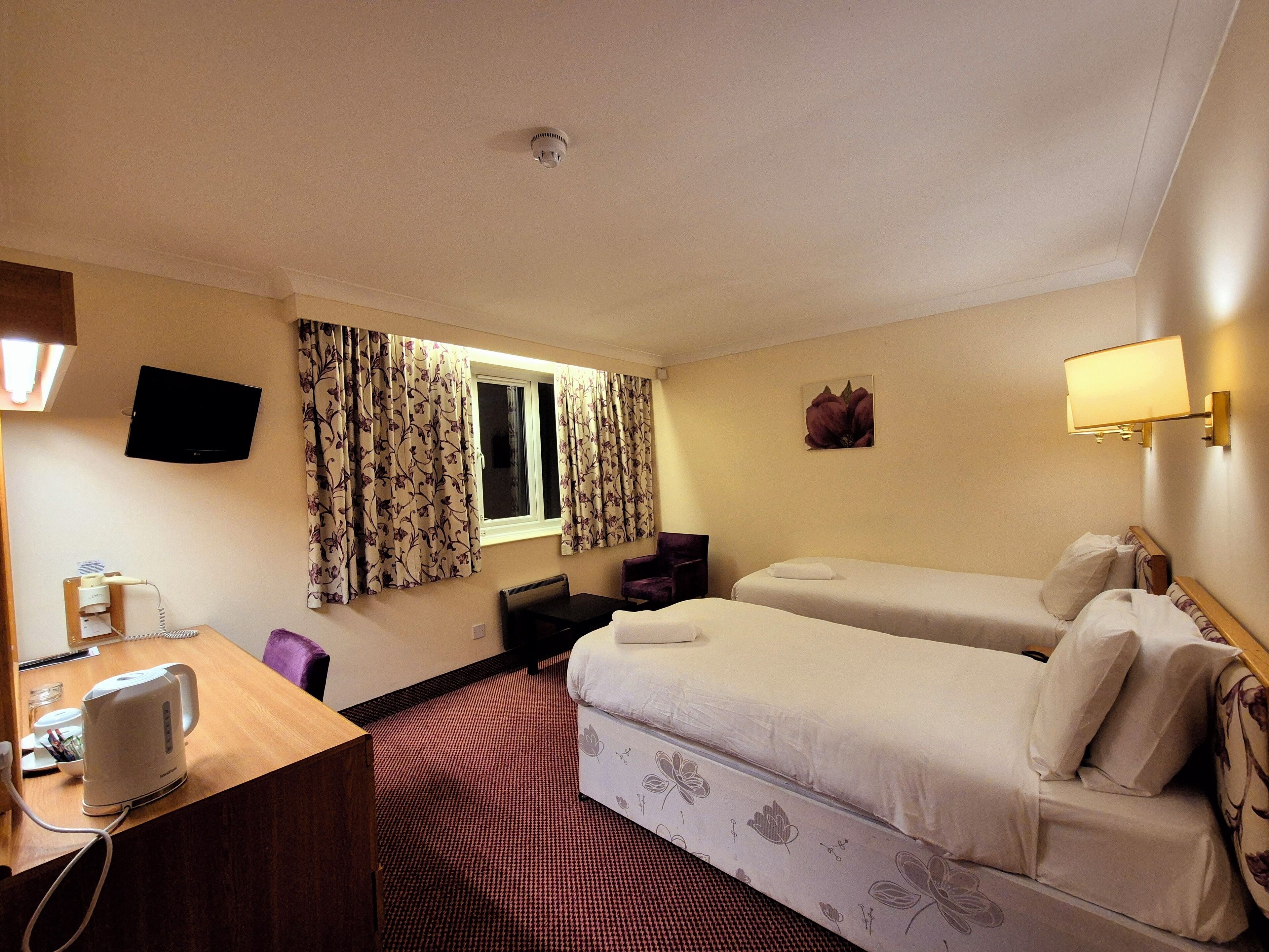 A hotel room with two beds, a desk with a kettle, a TV, and a window with curtains.