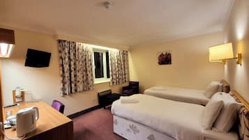 A hotel room with two beds, a desk with a kettle, a TV, and a window with curtains.