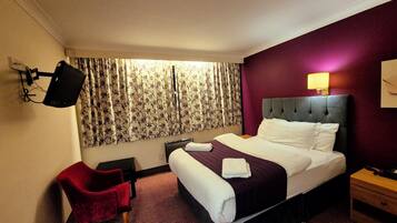 Une chambre d’hôtel avec un lit, un fauteuil rouge, un téléviseur fixé au mur et une fenêtre avec des rideaux.