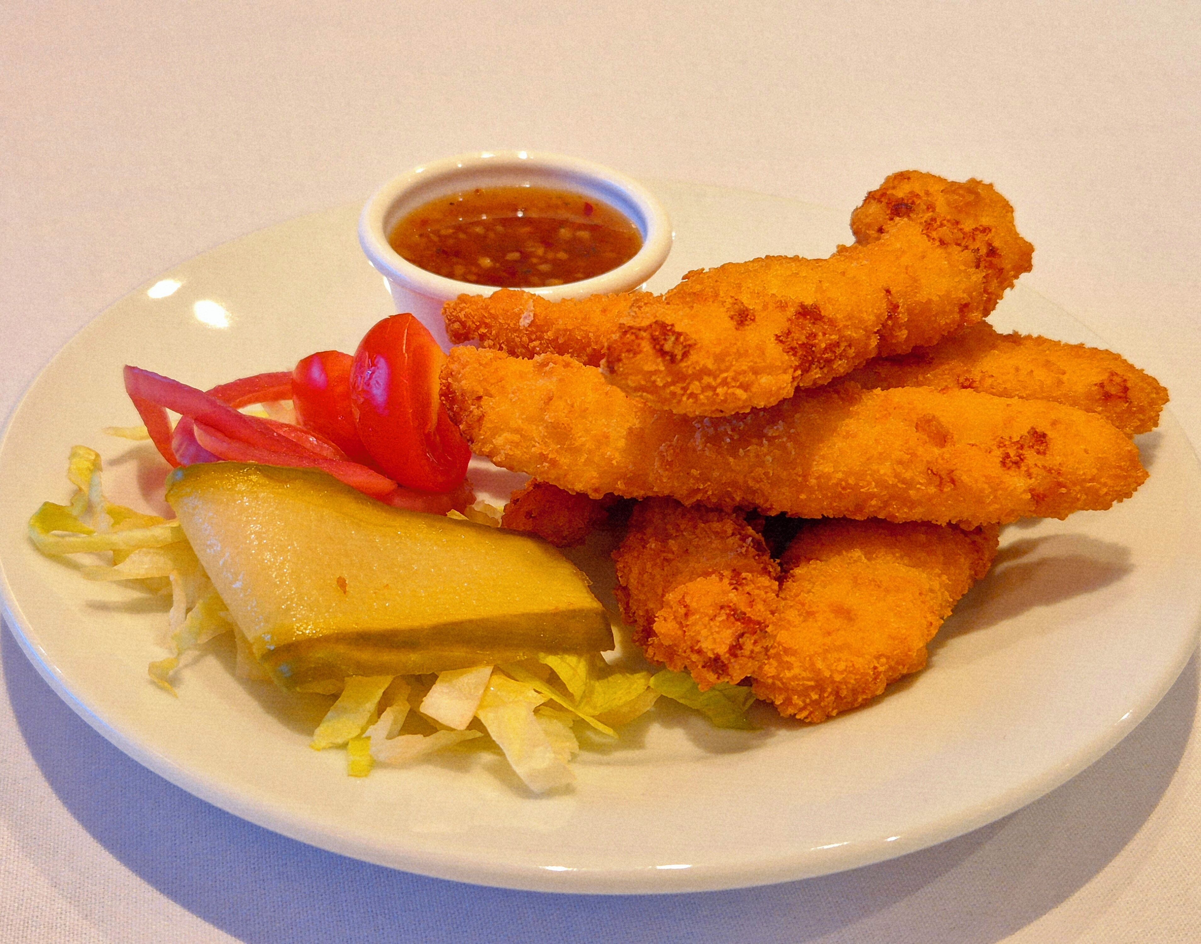 A plate of breaded and fried food items, possibly chicken tenders, accompanied by a small bowl of dipping sauce, a pickle, and some fresh vegetables.