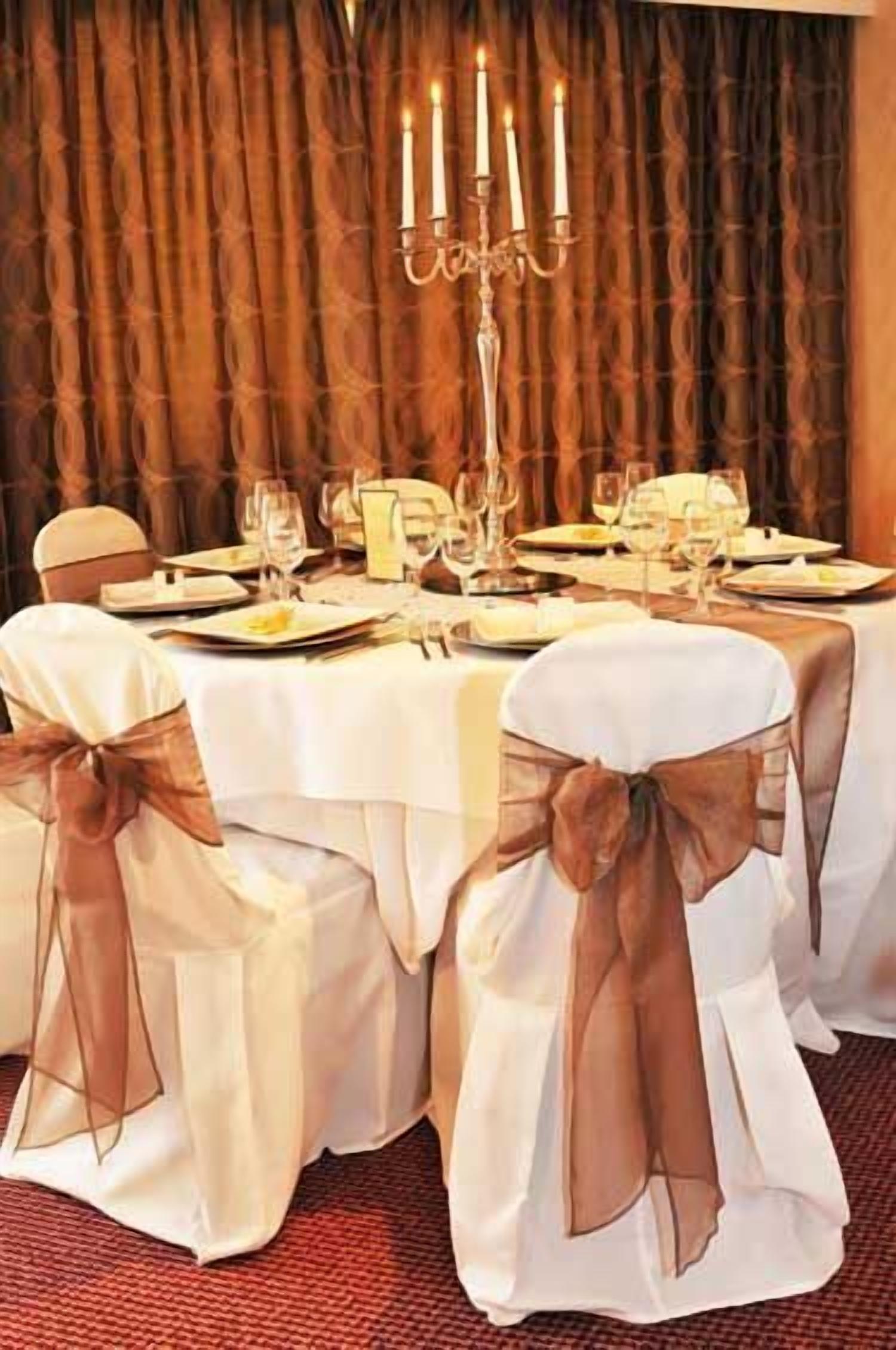 A formal dining table set for a meal with white tablecloths and brown sashes on chairs.