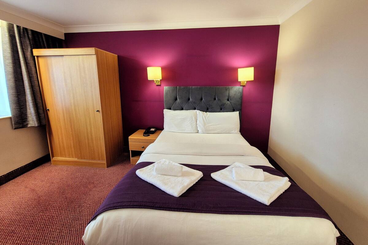 A hotel room with a bed, two bedside lamps, a nightstand, a wooden wardrobe, and a red carpet.