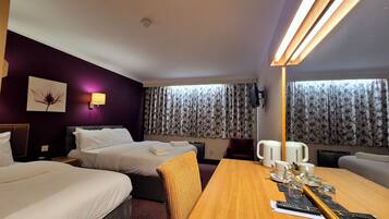 A hotel room with two beds, a wooden dining table with remote controls, a coffee maker, and a TV on the wall.